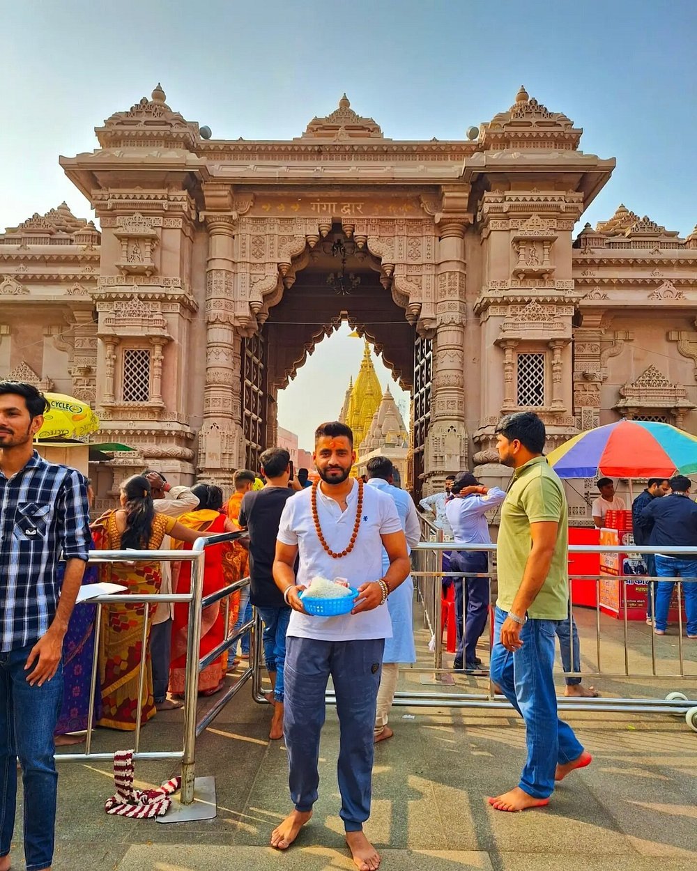 Man wearing a white shirt and beaded necklace stands barefoot, holding a blue basket, at an ornate temple gate with a crowd around him.